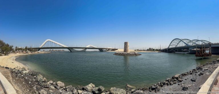 Wide view of Abu Dhabi’s Sheikh Zayed Bridge with its repeating white arches spanning behind the cylindrical Al Maqta Watchtower, rising from a stone base in calm reflective water.