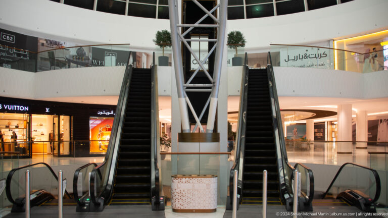 Frontal view of two parallel black escalators ascending between shops in a bright Abu Dhabi shopping mall interior with Arabic and English retail signage.