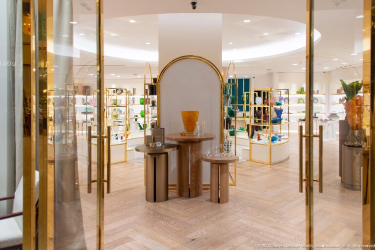 Frontal view into a glass-walled luxury boutique in an Abu Dhabi mall, with a minimalist white interior, gold accents, and a central display pedestal.