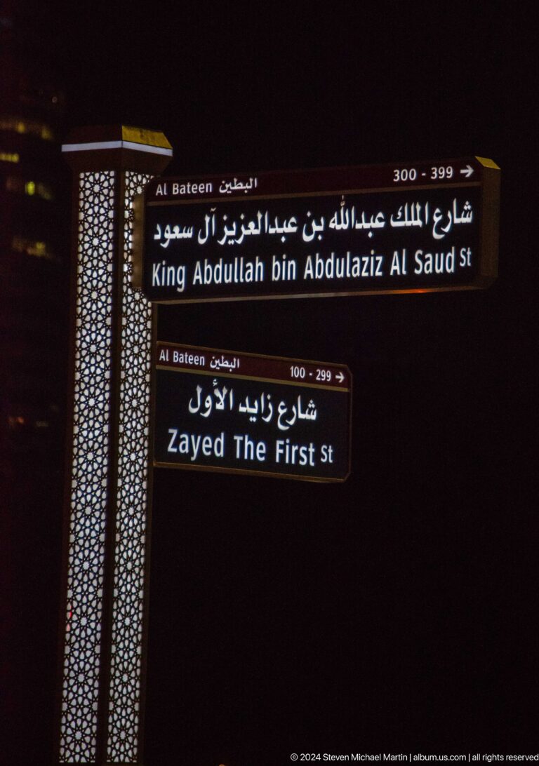 Nighttime view of bilingual Arabic-English street signs indicating directions to Imam Al-Tayeb Mosque and Zayed The First Street in Abu Dhabi.
