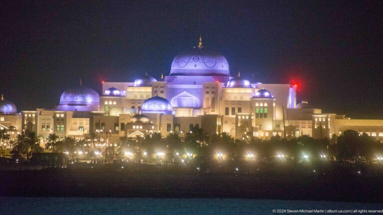 Distant nighttime view of Emirates Palace Mandarin Oriental in Abu Dhabi, its blue-violet central dome and long, warmly lit facade shining above a dark foreground.
