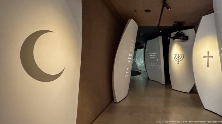 Contemporary gallery corridor at the Abrahamic Family House with a large black crescent on the left wall and a menorah symbol on a curved white panel ahead.
