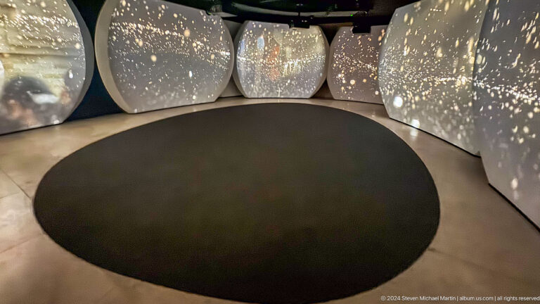 Low-lit interactive installation at the Abrahamic Family House with a circular dark floor surrounded by faceted seating modules dotted with small star-like lights.