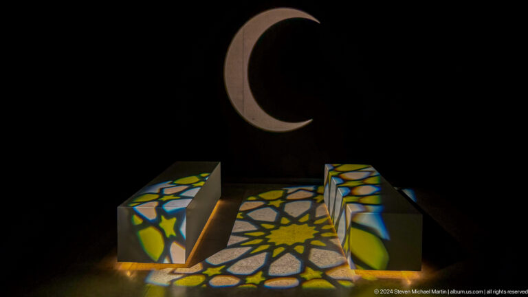 Dark gallery at the Abrahamic Family House with a large projected crescent above three low blocks illuminated by colorful Islamic geometric star patterns.