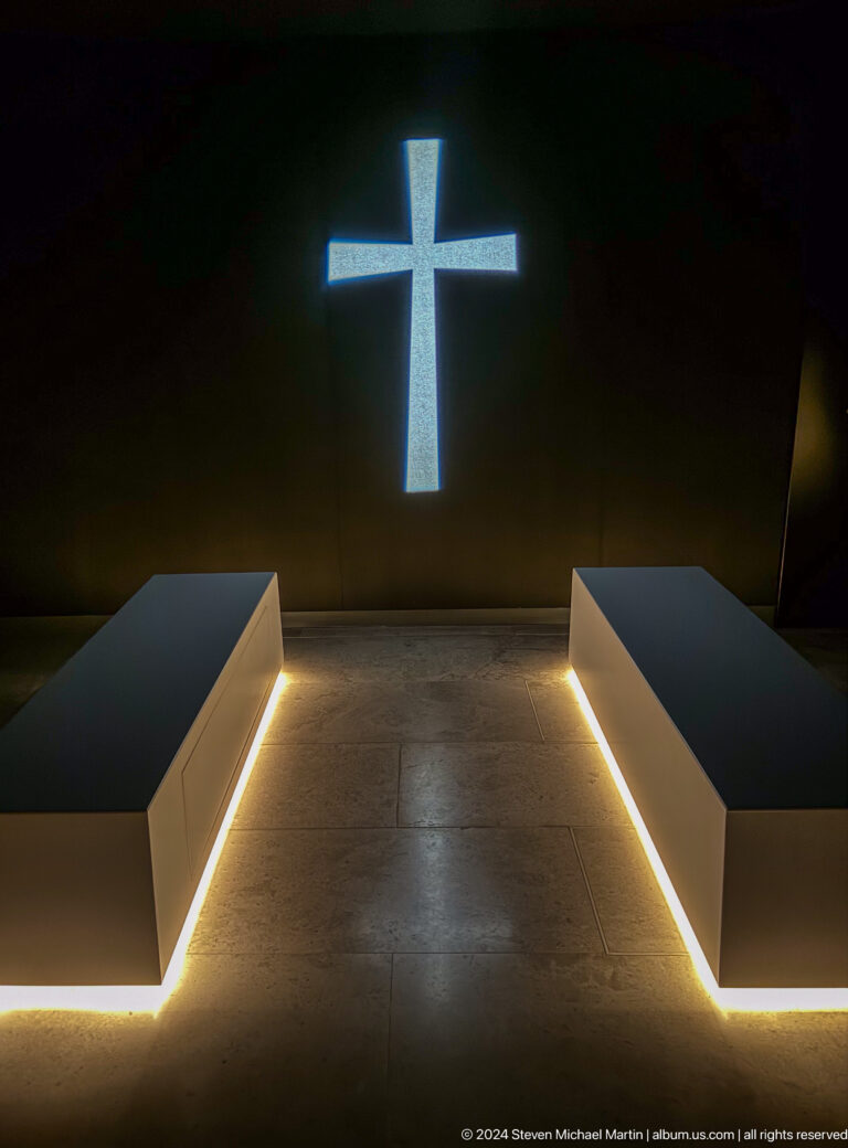 Dim interior reflection space at the Abrahamic Family House with two low parallel benches outlined by warm strip lighting, leading toward a narrow vertical opening of bright light at the far wall.