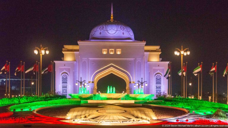 Frontal night view of the domed entrance gateway at Qasr Al Watan, lit in blue and green and reflected in a long pool lined with lights.