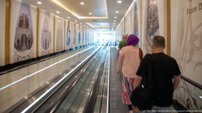 Two visitors walking away through a long moving-walkway corridor with wall displays.