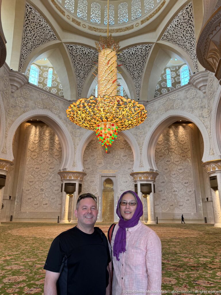 Smiling man and woman in a purple headscarf beneath a large chandelier and carved marble arches.