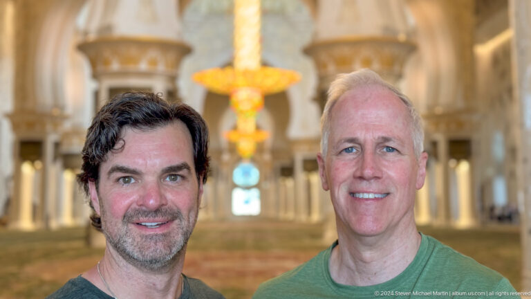 Close portrait of two smiling men with a chandelier and mosque interior blurred behind them.