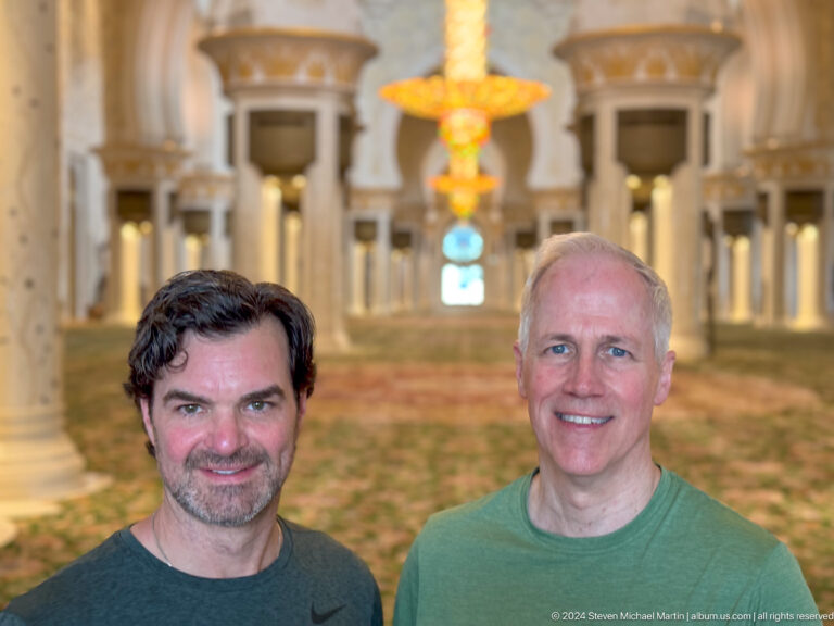 Two men pose in front of a floral prayer carpet, columns, arches, and a blurred chandelier.