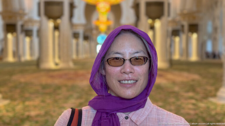 Smiling woman in glasses and a purple headscarf with a mosque chandelier blurred behind her.