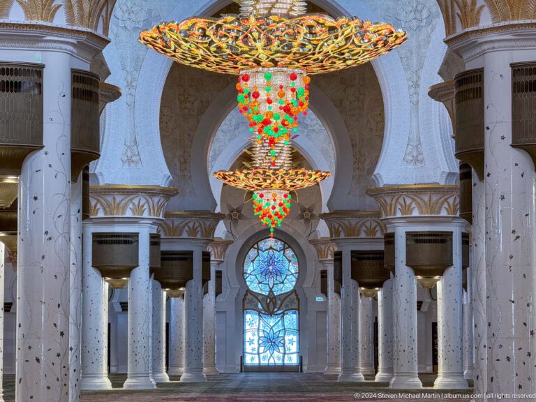 Two ornate chandeliers align above a blue stained-glass doorway between white marble columns and carved arches.