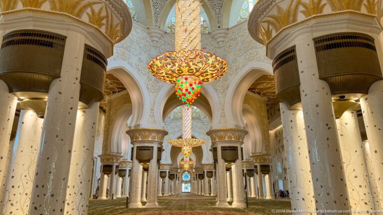 Two gold chandeliers aligned above blue stained glass between white marble columns and arches.