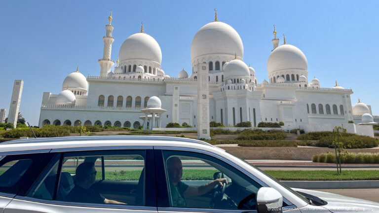 Passing car in front of the mosque’s white domes, arcade, and minaret.