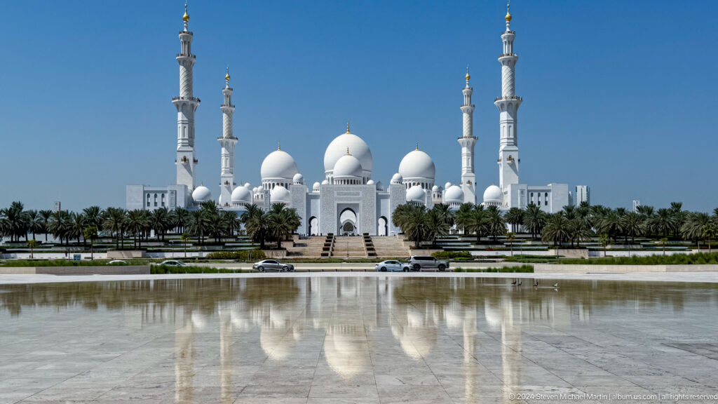 Wide view of white mosque domes and minarets reflected in a long pool.