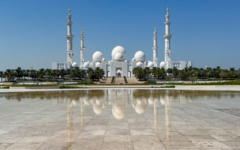 Centered mosque with white domes and four minarets reflected in a broad pool.