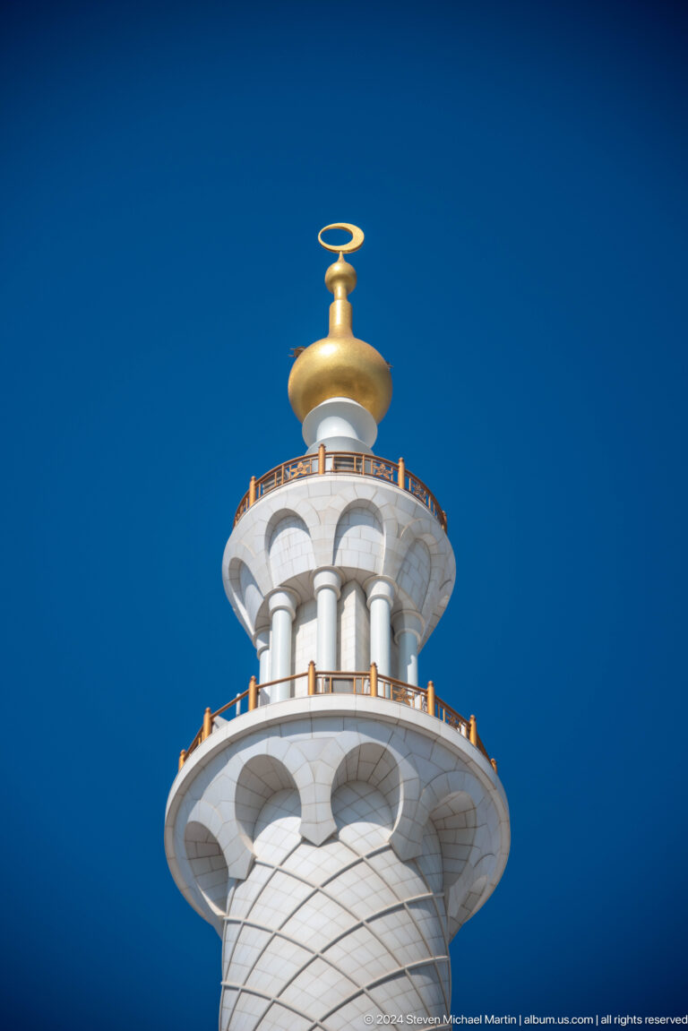 Upper minaret with white columns, blue band, and gold bulb finial against blue sky.