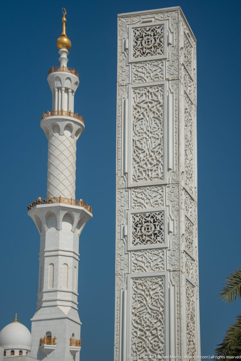 Carved white minaret shaft with geometric relief panels and a projecting balcony.