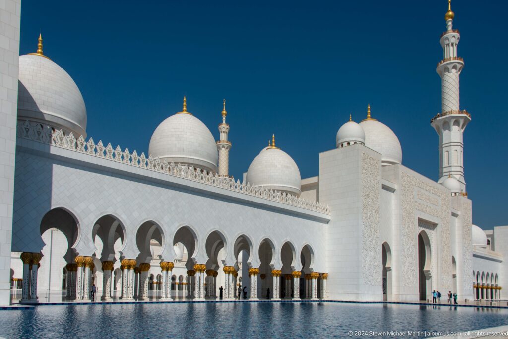 White mosque arcade with gold capitals, domes, and a minaret under blue sky.
