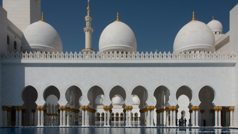 White domes and a minaret above a repeated arcade with gold capitals.