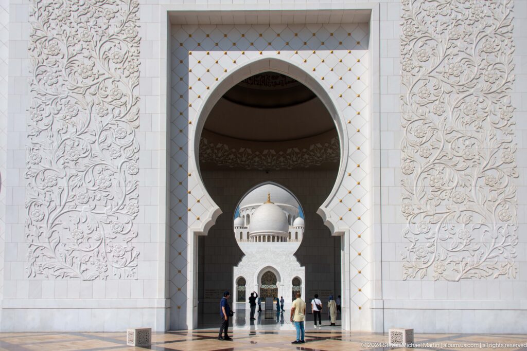Visitors beneath a large carved white entrance arch with a dome visible beyond.
