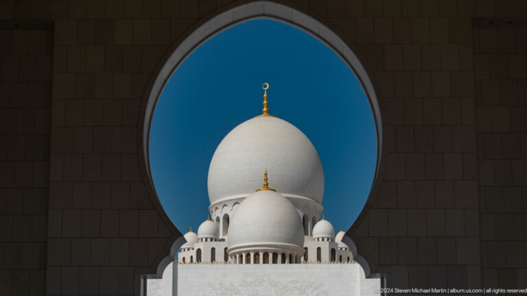 White mosque dome centered within a dark circular opening against blue sky.
