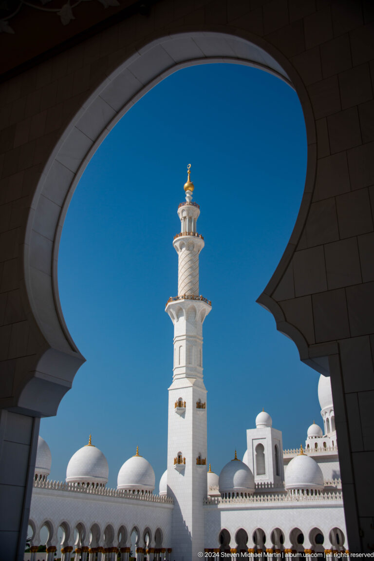 White minaret centered between dark scalloped arch edges against blue sky.