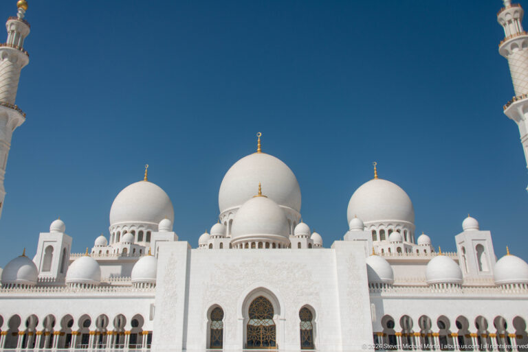 Three white domes with gold finials above a symmetrical arched façade.