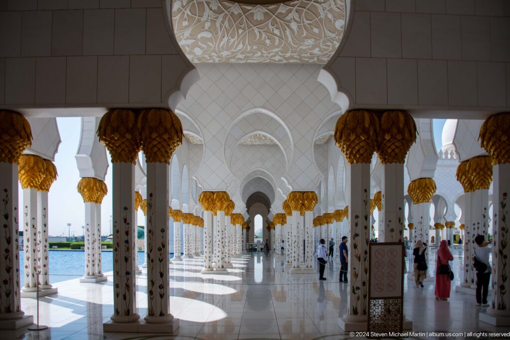 Long white mosque colonnade with gold capitals and visitors in the distance.