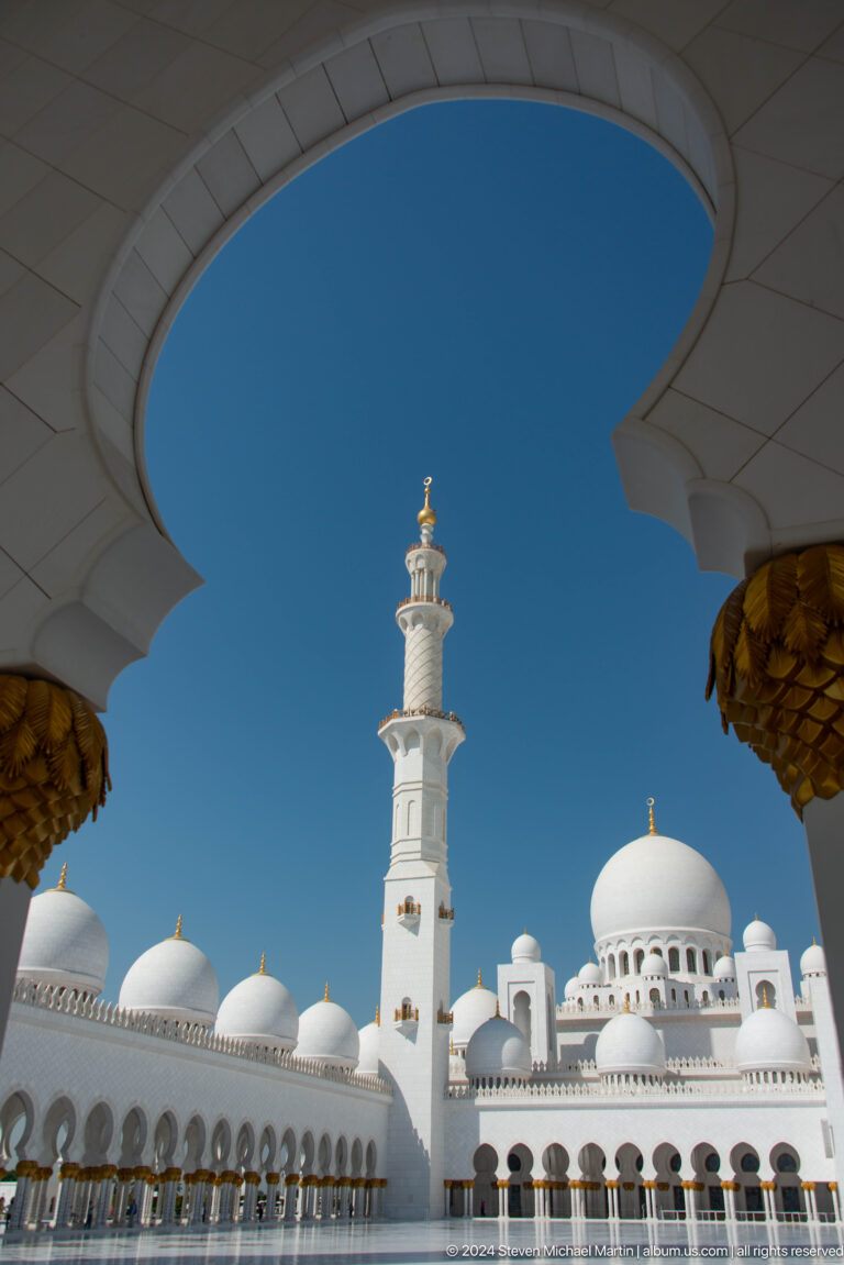 White minaret framed by dark scalloped arches with a small dome below.