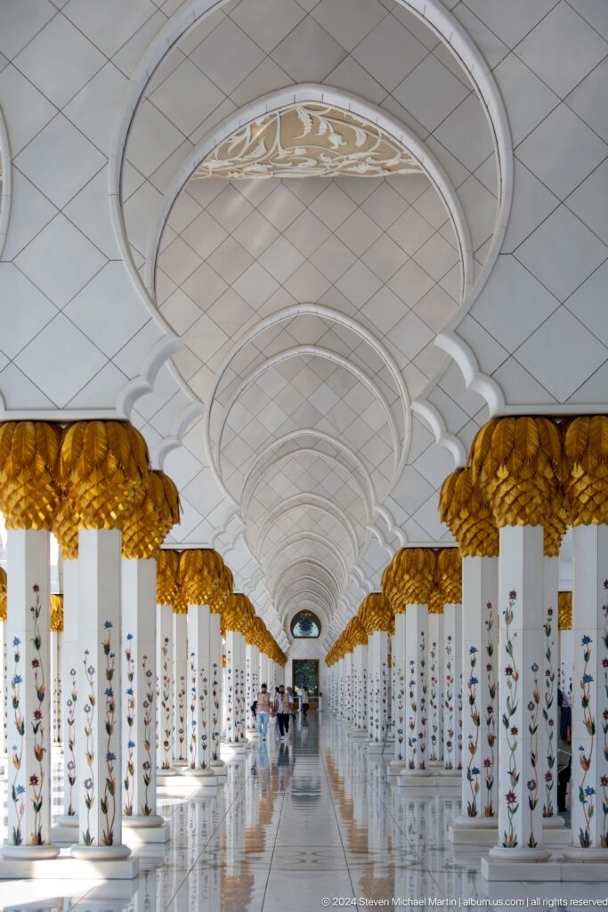 Symmetrical white arcade lined with repeating gold capitals and a bright vanishing point.