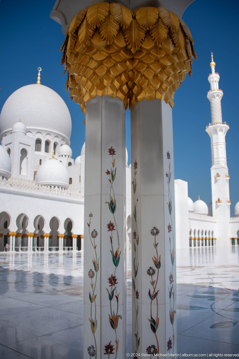 Floral-inlaid column with gold capital framing white arcades, domes, and a minaret.
