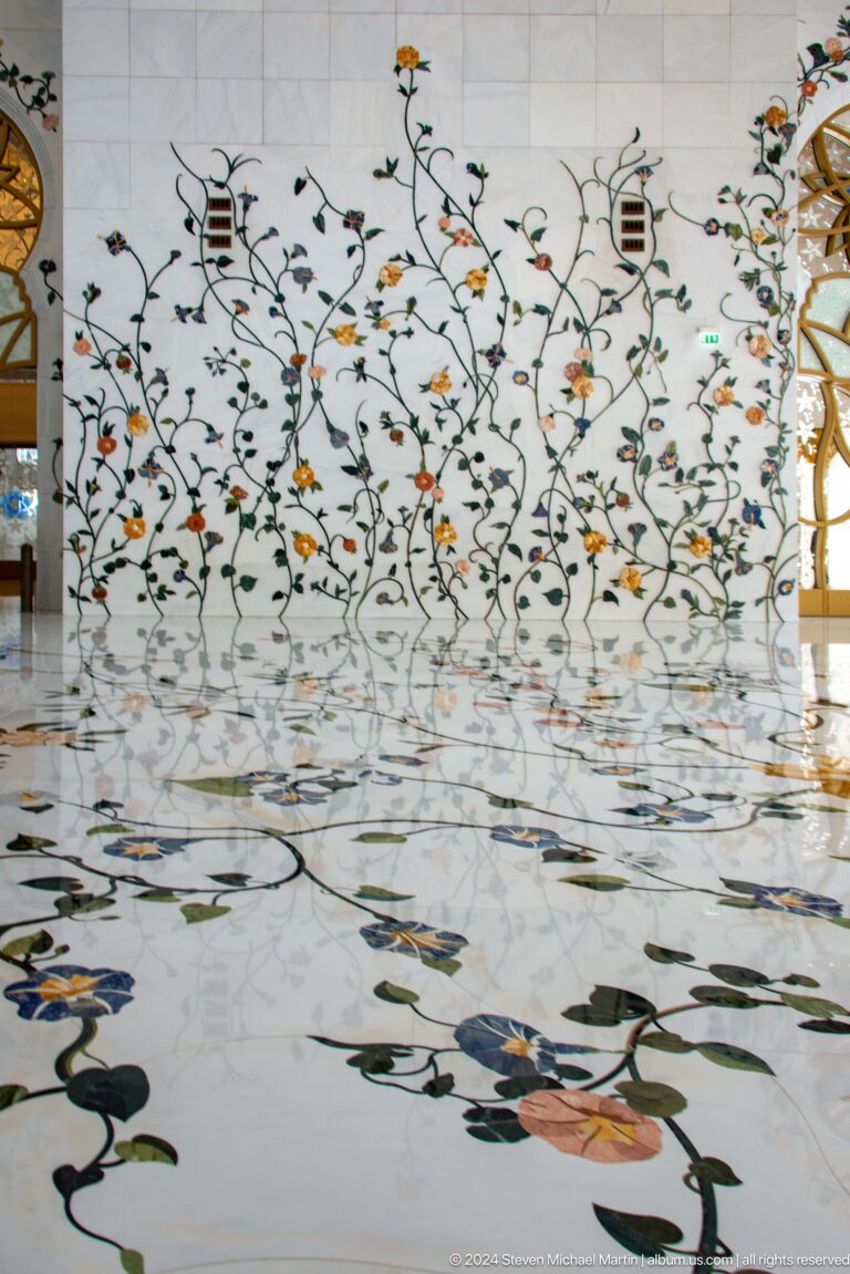Multicolored floral vines inlaid in a white marble wall reflect across a polished marble floor.