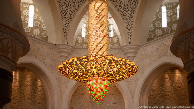 Large gilded chandelier with red, green, and amber glass drops beneath pointed arches.