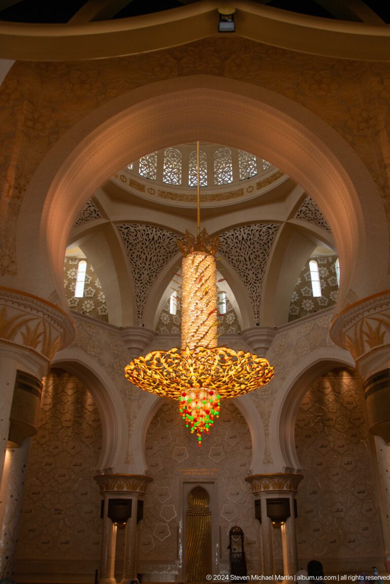 Upward view of a chandelier framed by ribbed arches, carved screens, and narrow windows.