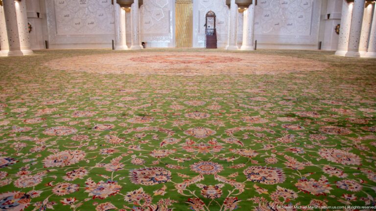 Low view across a green floral prayer carpet toward the decorated prayer-hall wall.