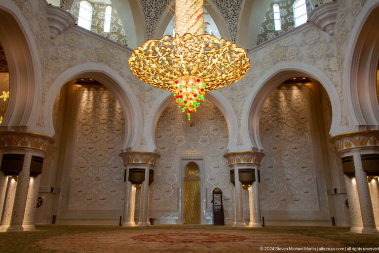 Symmetrical prayer hall with a monumental chandelier, pointed arches, white columns, and green carpet.