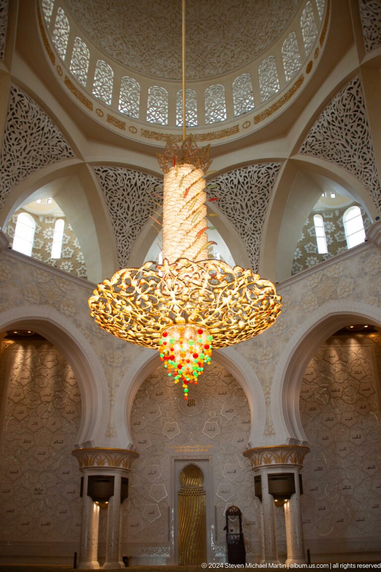 Close view of a gilded chandelier with colored glass drops between pale pointed arches.