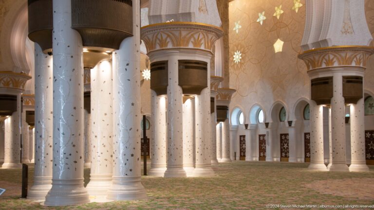 Rows of white marble columns with dark gold-trimmed capitals rise above a green floral carpet.