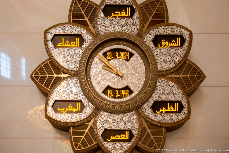 Decorative flower-shaped prayer-time clock with Arabic labels, analog hands, and digital date and time.