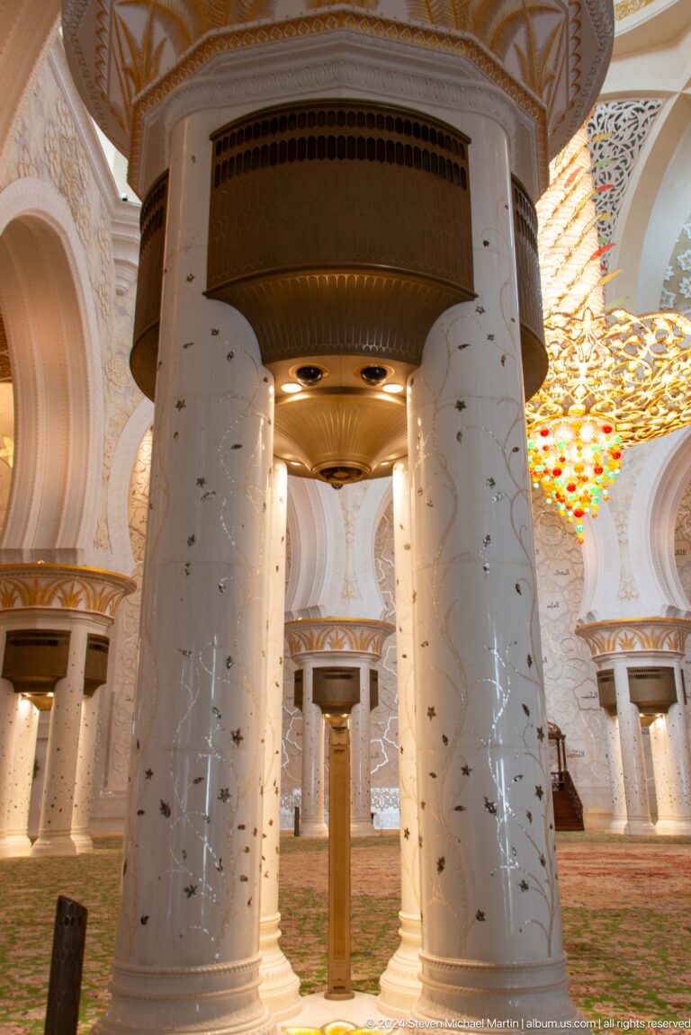 White marble columns with fine floral inlay and broad gold-toned capitals beneath ceiling lights.