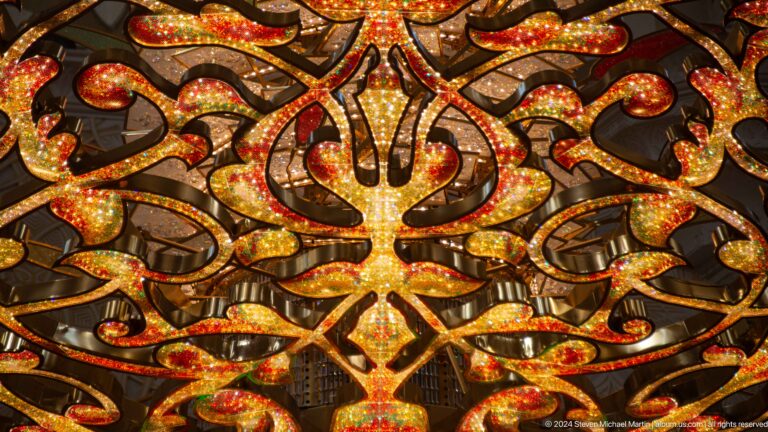 Extreme close-up of illuminated gilded arabesque chandelier scrollwork with red and green glass accents.