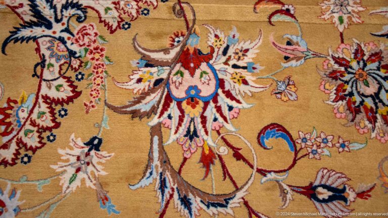 Close-up of a beige prayer carpet with a large red, blue, white, and green floral motif.