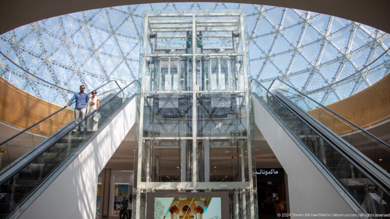 Glass elevator centered between two escalators under a geometric roof.