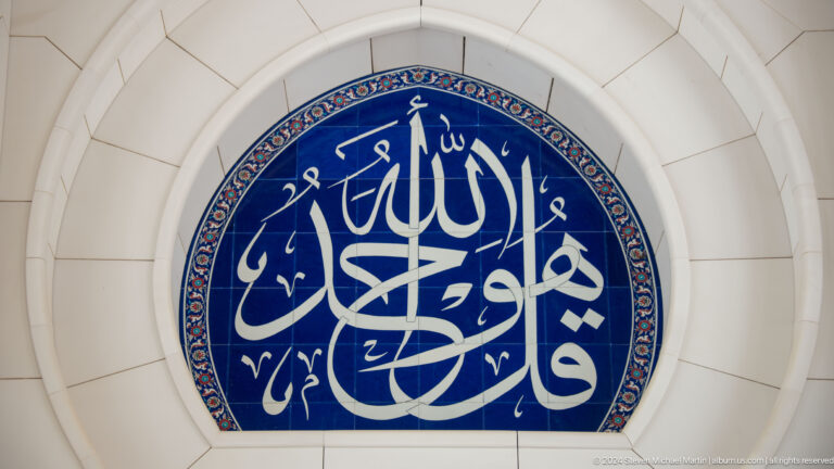 White Arabic calligraphy on a dark-blue oval panel set within a pale arched wall.