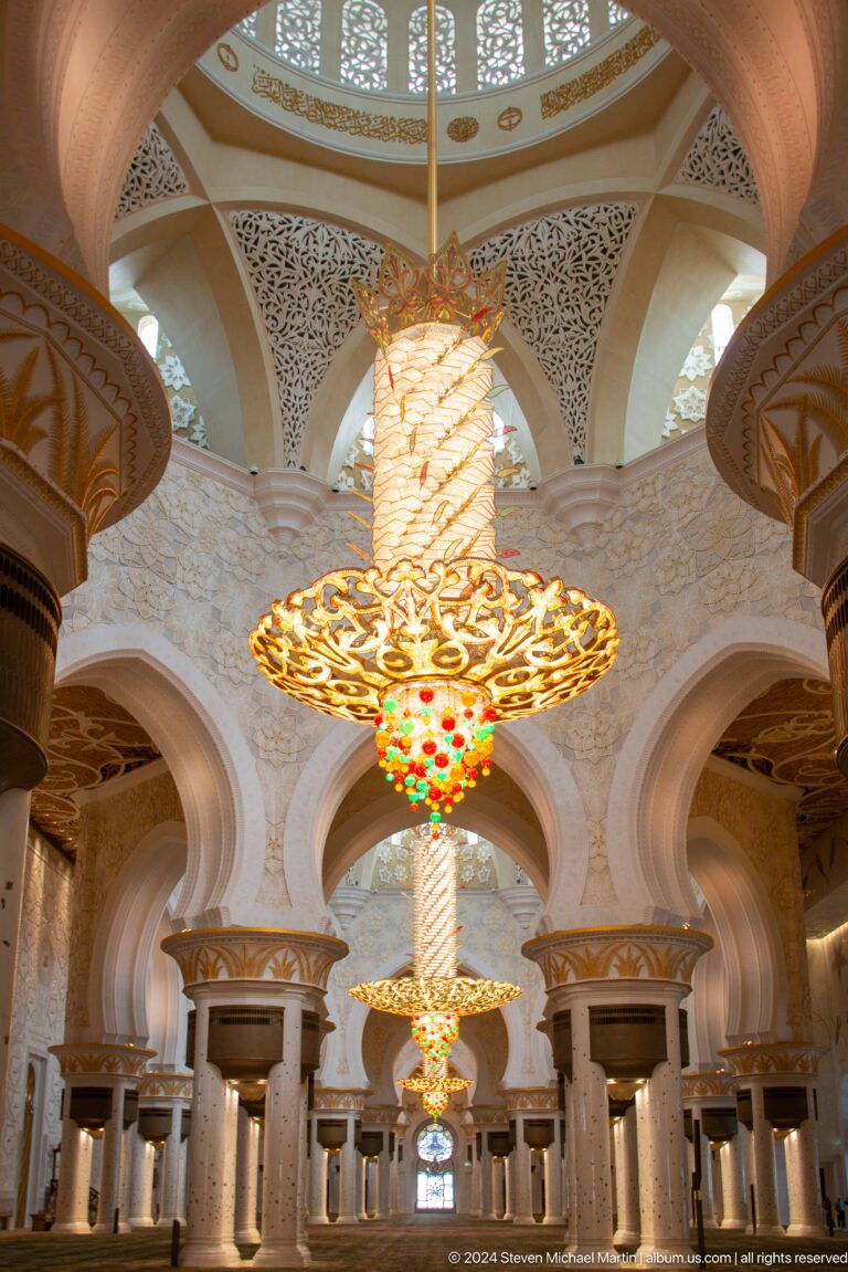 Large gilded chandelier with colored glass drops framed by pale vaulted arches.