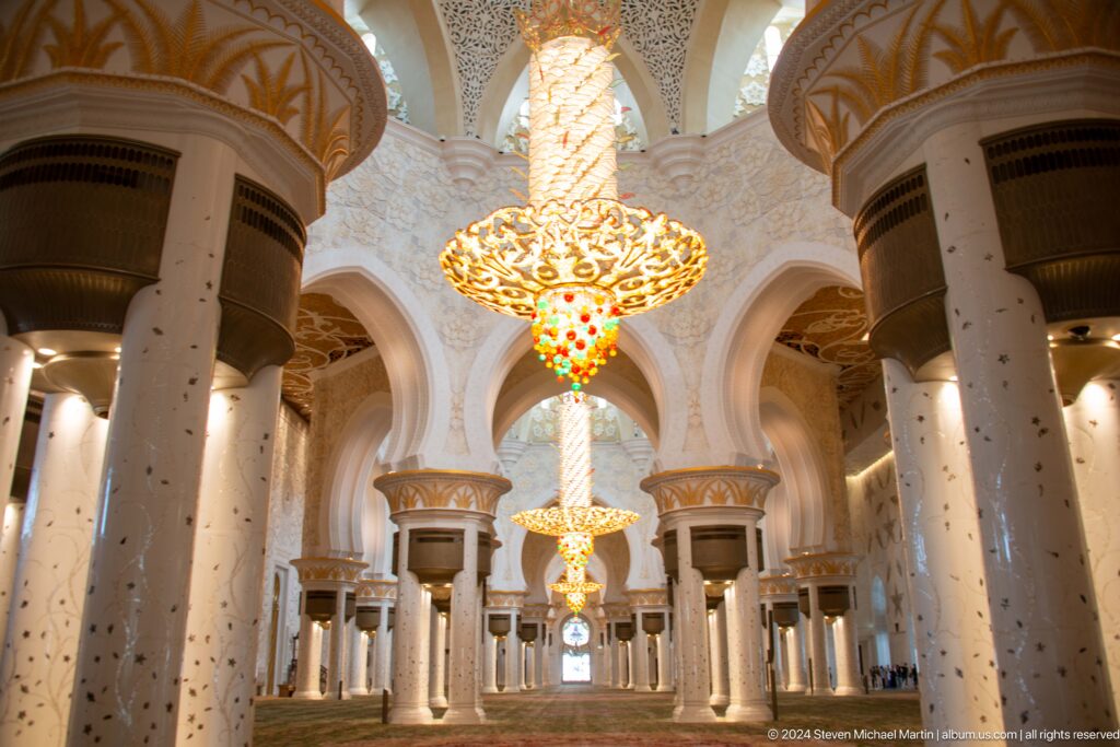 Symmetrical prayer hall with chandelier, pointed arches, white columns, gold capitals, and green carpet.