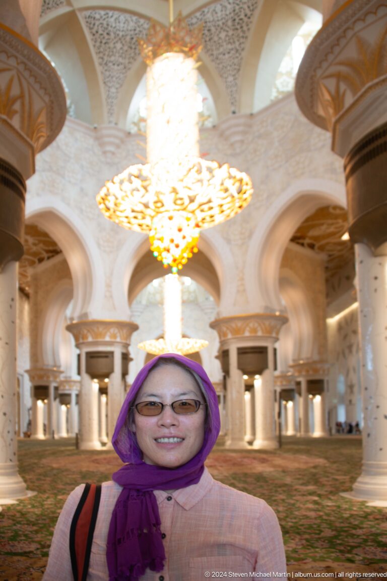 Smiling visitor in a purple headscarf beneath a glowing chandelier inside the mosque.