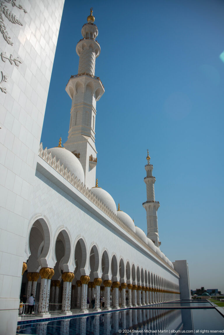 White mosque arcade and low dome with a distant minaret against blue sky.