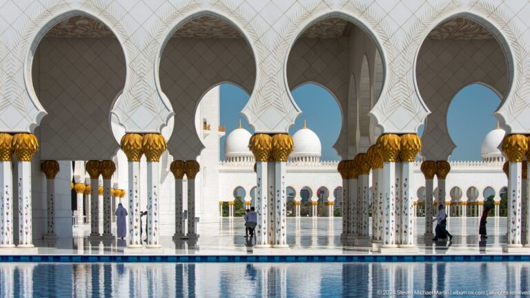 Repeated scalloped arches and gold-topped columns frame visitors crossing the reflective courtyard.